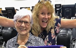 A staff member with arms wide stands behind a member on the wellness floor. Background includes several cardio machines.