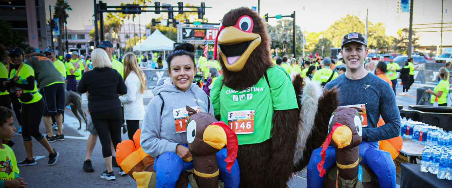 Tampa YMCA Turkey Gobble mascot poses in the middle of two people. The attendees are wearing turkey costumes that look like they're riding turkeys. Background: many event participants socializing, wearing neon yellow race shirts, and tables of many water bottles on the right.