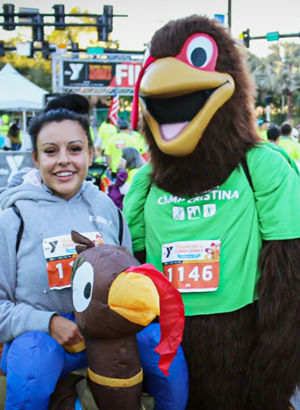 Tampa YMCA Turkey Gobble mascot poses in the middle of two people. The attendees are wearing turkey costumes that look like they're riding turkeys. Background: many event participants socializing, wearing neon yellow race shirts, and tables of many water bottles on the right.