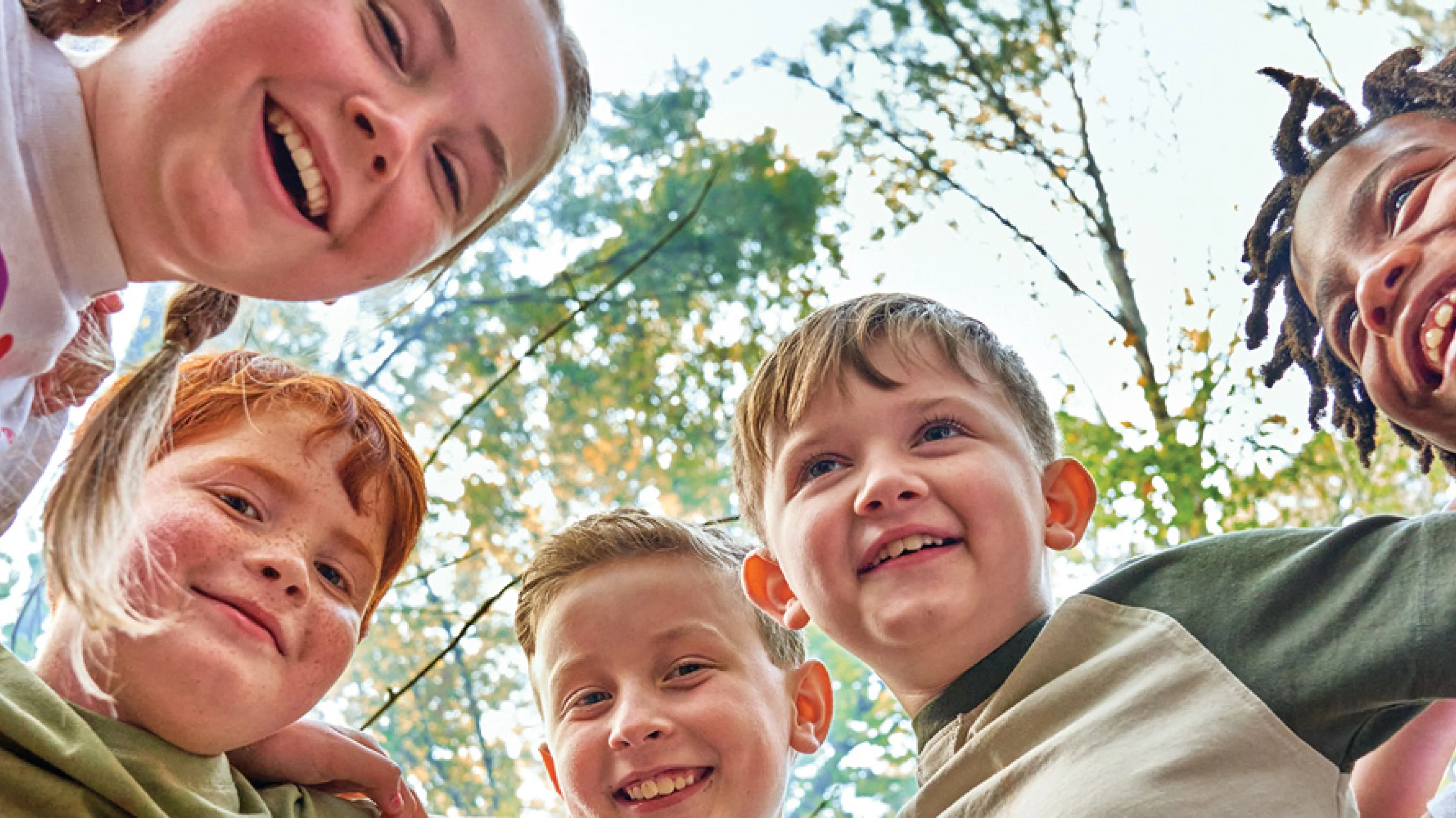 Five kids huddling, smiling, looking down at the camera, lens is pointing up at their faces. Trees background of the photo.