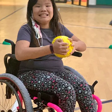 Young girl smiling in wheelchair, holding yellow ball in basketball gym